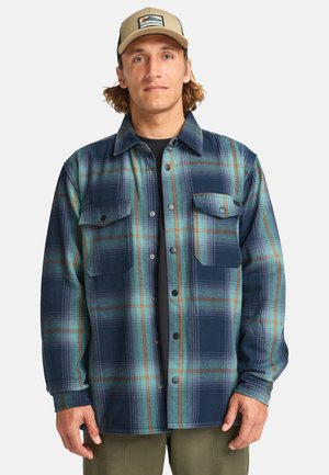 Man wearing a beige cap and blue-green plaid button-up shirt with two chest pockets over a black shirt.
