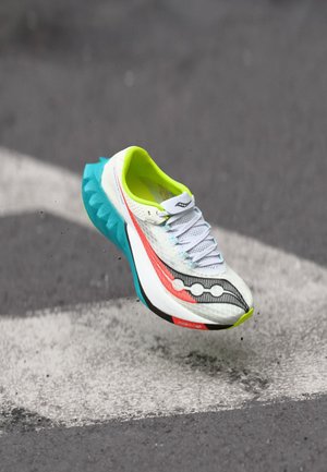 White running shoe with red and black stripes, neon yellow lining, and blue sole mid-air above gray asphalt with white arrow marking.