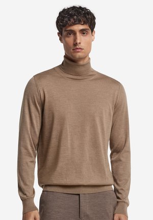 Mann, der einen hellbraunen Rollkragenpullover und passende Hose trägt, steht vor einem schlichten weißen Hintergrund.