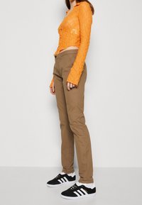 Pomarańczowa koronkowa krótka bluzka z długim rękawem i zapięciem na guziki, w połączeniu z beżowymi dopasowanymi spodniami oraz czarnymi zamszowymi sneakersami z białymi paskami.