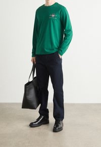 Zielony longsleeve z logo, granatowe spodnie, czarne skórzane buty oraz duża czarna torba typu tote na tle jasnoszarym.