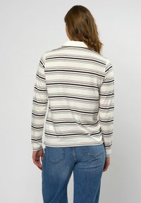 Vrouw die een crèmekleurig shirt met lange mouwen draagt, met zwarte en roze horizontale strepen, en een blauwe spijkerbroek, gezien van achteren.