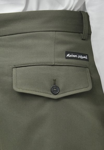 Olive roheline püksid, mis sisaldavad nööbiga klappi taskut, nähtavat õmblemist ja musta sildi valge tekstiga, millel on kirjas "maison kitsuné."