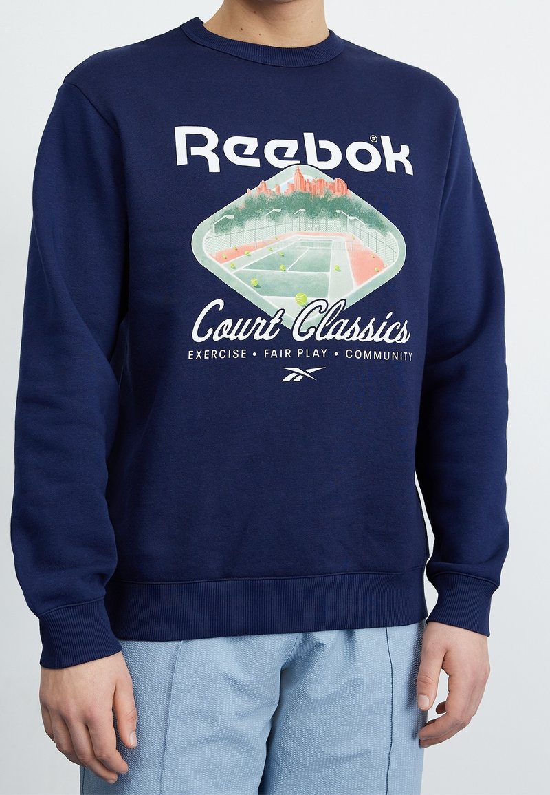 Mörkblå sweatshirt med ett grafiskt motiv av en tennisbana och Reebok-logotypen. Tyget är mjukt, med ribbade muddar och nederkant.