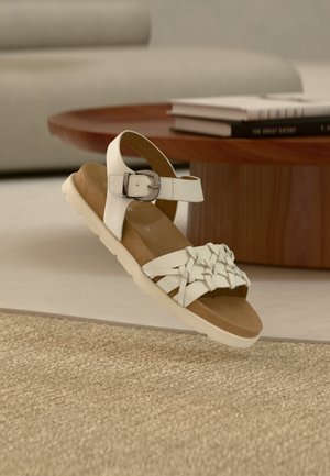 White sandal featuring a woven strap design and adjustable ankle buckle. The sole is beige with a textured surface for grip.