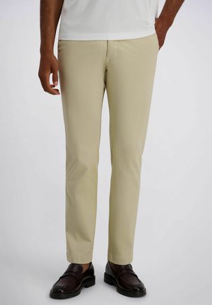 Hombre que lleva pantalones beige de pierna recta y mocasines de cuero marrón oscuro, de pie con una mano en el bolsillo contra un fondo liso.