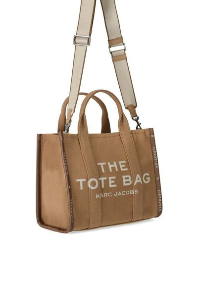 Beige kangaskassi, jossa on kaksinkertaiset kahvat ja säädettävä olkahihna, etupuolella lukee "THE TOTE BAG MARC JACOBS".