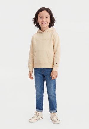 Enfant souriant aux cheveux mi-longs, portant un sweat à capuche beige Champion, un jean bleu retroussé et des baskets montantes beiges, debout sur fond blanc.