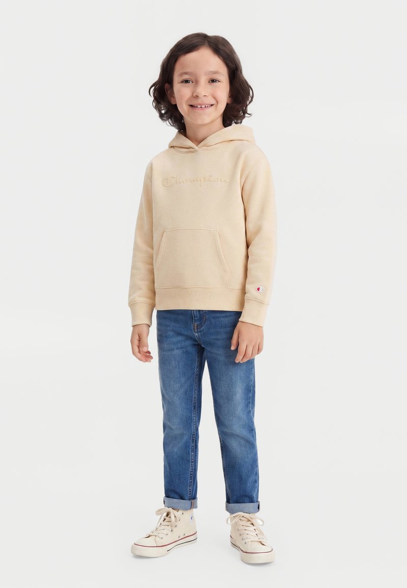 Enfant souriant aux cheveux mi-longs, portant un sweat à capuche beige Champion, un jean bleu retroussé et des baskets montantes beiges, debout sur fond blanc.