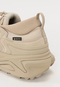 Tenis bej cu un material texturat și talpă netedă. Dispune de eticheta Gore-Tex, guler moale la gleznă și toc rotunjit, având un design ușor.