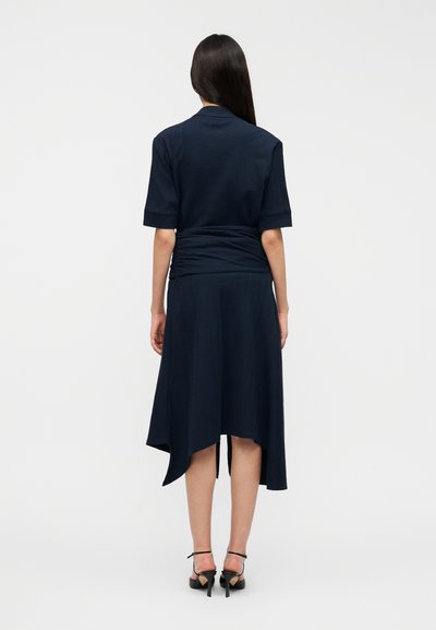 Μπλε ναυτικό φόρεμα με κοντά μανίκια, υφασμάτινη υφή, καμπυλωτό τελείωμα και λεπτομέρεια με σούρες στη μέση. Φορεμένο με μαύρα σανδάλια με λουράκια.