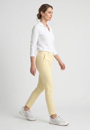 Kvinde går, iført hvid langærmet polo, lysegule smalle bukser og hvide snøresneakers med teksturerede såler.