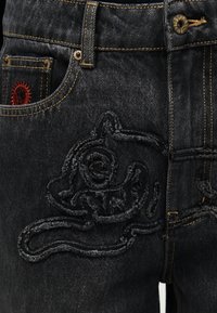 Μαύρο τζιν από ντένιμ με παχύ μαύρο κεντητό σχέδιο γάτας στο μηρό και κόκκινο έμβλημα ήλιου μέσα στην μπροστινή τσεπούλα, με κίτρινες ραφές λεπτομέρειες.