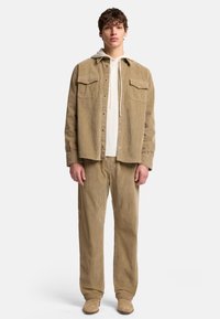 Beige Kordjacke mit zwei Brusttaschen, übereinander getragen mit einem weißen Hoodie und passenden Kordhosen, kombiniert mit beige Farbdämpfern.