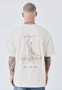 Crème t-shirt met korte mouwen, featuring een grijze afbeelding van het Vrijheidsbeeld en tekst op de achterkant, vervaardigd uit zachte, lichtgewicht stof.