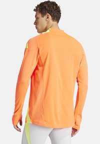 Langarmshirt van feloranje stof met textuurdetails, met reflecterende accenten op de schouders en zoom voor zichtbaarheid.