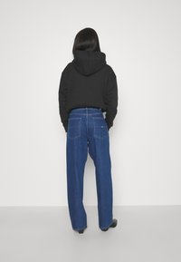 Person med langt mørkt hår iført en svart hettegenser og blå rette jeans, stående vendt mot en enkel hvit vegg.