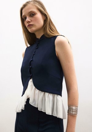 Młoda kobieta ubrana w granatową kamizelkę bez rękawów z przednimi guzikami na białej bluzce z falbankami, ozdobiona licznymi srebrnymi bransoletkami.