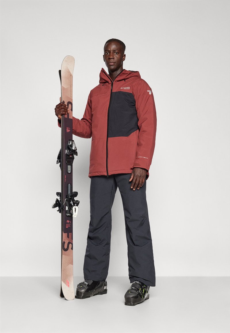 Abbigliamento da sci composto da un giaccone isolato color marrone scuro e nero, pantaloni da sci scuri e sci con attacchi, impostato su uno sfondo semplice.