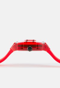 Montre en plastique rouge avec un boîtier transparent, un cadran rond avec des accents noirs et un strap lisse et flexible. Bouton situé sur le côté.