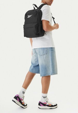 Persona con camiseta blanca, pantalones cortos de mezclilla clara, calcetines blancos de Reebok, zapatillas de colores y mochila negra con un logo blanco.