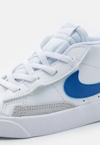 Vita sneakers med en blå Nike swoosh, grå läderdetaljer och vita snören. Texturerad gummisula för grepp och stabilitet.