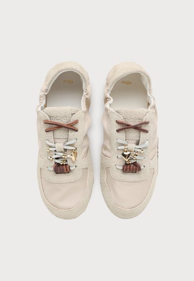 Paire de baskets beige en daim et cuir avec des lacets marron et blancs, décorées de breloques dorées et de pendentifs en forme de cœur.
