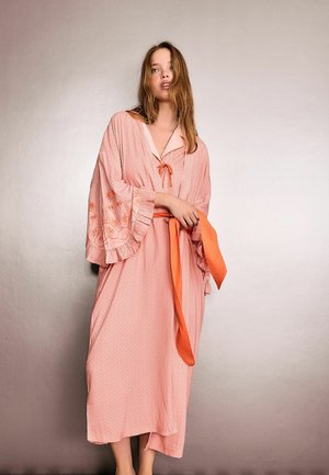 Nő hosszú rózsaszín ruhában, apró mintákkal, széles fodros ujjakkal, virágmintás díszítéssel, és narancssárga övvel a kezében.