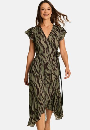 Femme marchant, vêtue d'une robe portefeuille à imprimé animal vert et noir, avec des manches volantées et un ourlet asymétrique, sur un fond uni.