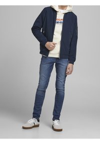 Marynarska, lekka kurtka bomber, kremowa bluza z kapturem z nadrukiem graficznym, niebieskie slim-fit dżinsy i białe trampki z czarnymi i brązowymi akcentami.