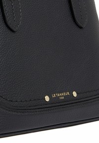 Primo piano di una borsa a tracolla in pelle nera texturizzata con borchie dorate e dettaglio del logo "Le Tanneur 1898" in oro vicino al bordo inferiore.