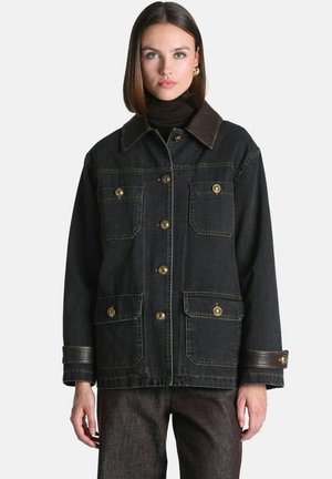 Femme aux cheveux lisses mi-longs portant une veste en jean foncée avec des poches boutonnées et un col et des poignets en cuir.