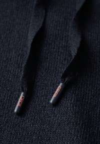 Marinblått stickat tyg med två mörka dragsnören med svarta ändar, var och en märkt med 'CECIL' i orange text.