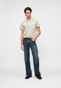 Fiatal férfi áll, bézs pólót, barna övet, kék egyenes lábú farmernadrágot és fekete cipőt visel egy sima fehér háttér előtt.