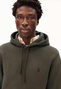Hoodie verde scuro con una tasca a marsupio frontale, cappuccio regolabile con cordino e piccolo logo sul petto. Texture morbida e vestibilità rilassata sopra una camicia con colletto.