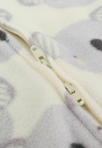 Kombinezon z polaru o kremowej podstawie, z jasnofioletowymi i szarymi wzorami zwierzęcymi oraz plastikowym zamkiem błyskawicznym z przodu. Miękka tekstura.