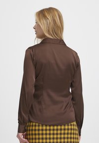 Braune Satin-Hemdbluse mit langen Ärmeln und Kragen. Der Rücken zeigt ein tailliertes Design, kombiniert mit einem gelb-schwarz karrierten Rock.