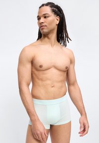 Μυώδης άντρας με κότσους dreadlocks, που φοράει ανοιχτό μπλε μποξεράκια, κοιτάζοντας προς τα πλάγια, στέκεται μπροστά σε λευκό φόντο.