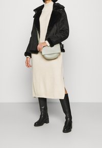 Zwarte nepbont gevoerde jas, crèmekleurige gebreide jurk, zwarte kniehoge laarzen en een groene schoudertas met een ronde vorm. Gladde texturen zichtbaar.