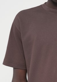 Rjava majica s kratkimi rokavi, narejena iz mehke bombažne tkanine, z okroglim izrezom in sproščenim krojem. Prikazuje gladko teksturo s čistimi šivi.