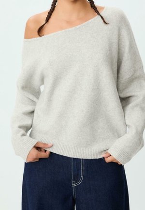 Woman wearing light gray off-shoulder knit sweater with long sleeves and dark blue high-waisted jeans, hands tucked in front pockets.