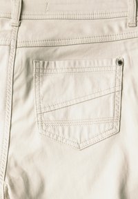 Pantalones de mezclilla beige que presentan un bolsillo trasero con costura diagonal, un acento de botón de metal y una textura suave. Diseño sencillo y discreto.
