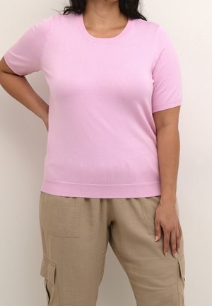Woman wearing a short-sleeve, light pink knit top and beige cargo pants, standing with one hand on hip and the other relaxed.