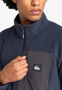 Giacca in pile blu marina con colletto alto, chiusura con zip e taschino grigio sul petto con logo. Il tessuto appare morbido e testurizzato.