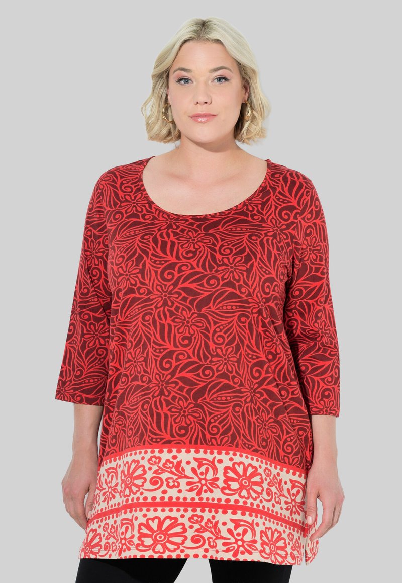 Túnica de patrón floral rojo con mangas tres cuartos. Presenta una sección superior sólida y un dobladillo detallado en un tono más claro. Tejido suave.