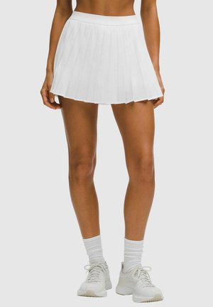 Tanned legs wearing a white pleated tennis skirt, white crew socks, and white athletic sneakers against a plain background.