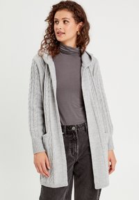 Lichtgrijze gebreide cardigan met een capuchon, voorzien van gestructureerde kabelpatronen, zijzakken en geribbelde manchetten. Draagt over een donkere top en broek.