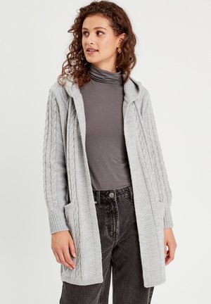Light gray knitted cardigan with a hood, featuring textured cable patterns, side pockets, and ribbed cuffs. Worn over a dark top and pants.