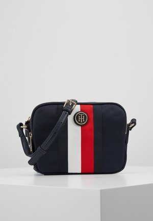 Sac bandoulière compact bleu marine avec bandoulière réglable, présentant des rayures verticales blanches, rouges et noires et un logo rond doré.