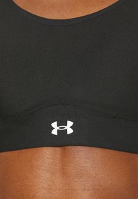 Melna sporta krūšturis ar gludu, elastīgu audumu. Aprīkots ar platu joslu un baltu Under Armour logo centrā. Apaļas kakla izgriezuma dizains.